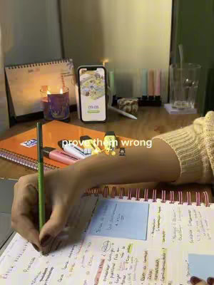A well-lit desk with study materials. A hand holding a pen over a notebook filled with notes is highlighted. A lit candle and a phone displaying a home screen are visible in the background. The overlay text reads 'prove them wrong'.