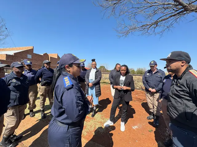 A group of people, including police officers in uniform, are standing in a circle outdoors under a sunny sky. The people are of diverse ethnicities and are engaged in what appears to be a discussion or meeting. Buildings and bare tree branches are visible in the background.