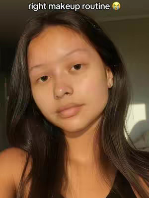 A selfie of a young woman with long dark hair and a neutral expression. She looks directly at the camera against a blurred background, possibly indoors with natural lighting.
