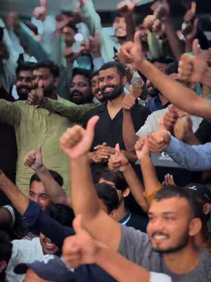A large crowd of people, primarily men, are raising their hands with thumbs up, with a man at the center of the image with a thumbs up.