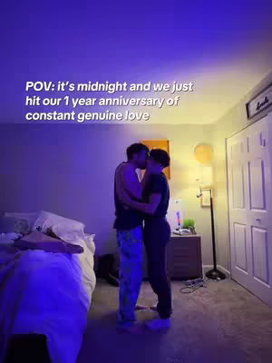 Two men kissing in a bedroom, lit by a blue light. A bed is in the foreground to the left, and the text overlay emphasizes their anniversary.