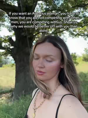 A woman with long hair stands outdoors with a blurred background of greenery and a tree, looking slightly upwards with closed eyes, wearing a black spaghetti strap top and a gold necklace.