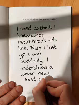 The image shows an open book on a wooden surface. The left page is visible, displaying handwritten text in black ink, likely a quote or a personal reflection. A pen is held in the bottom-right corner, with a hand writing on the page. The pages are white, creating contrast with the black text and the wooden surface. The composition is simple and focuses on the act of writing and the content within the book.
