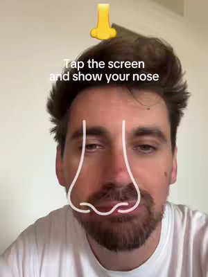 A man with dark hair and a light complexion is centered in the frame, wearing a white t-shirt. Above his head, a yellow nose emoji is placed. A white outline of a nose is drawn over his face. A text overlay reads, 'Tap the screen and show your nose.' The background is a plain, light-colored wall giving the impression of a simple, well-lit space.