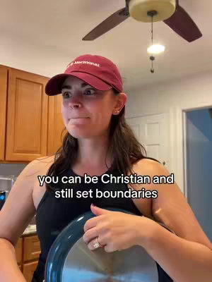 A woman in a maroon baseball cap and black tank top is holding the lid of a pan. She is in a kitchen with wood cabinets in the background.