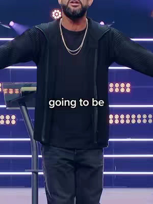 A man with a beard wearing a black jacket over a black shirt is standing in front of lights on a stage, arms outstretched with a message overlayed.