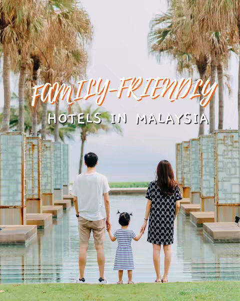 A photograph from a low angle, showing a family of three from behind. The parents are holding hands with a young child. They are standing on a grassy surface, in front of a body of water. The background includes a line of decorative structures, lined with tall palm trees and a light, overcast sky. The text overlay reads 'FAMILY-FRIENDLY HOTELS IN MALAYSIA' in handwritten-style script.