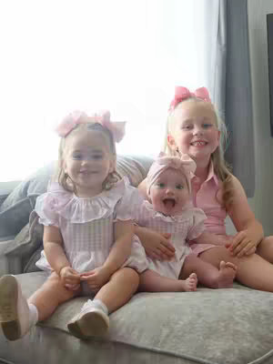 A photograph shows three young girls, all in a similar pink color scheme, sitting on a gray couch. The girl on the left has a large pink bow, a white top with a ruffled collar, and is looking at the camera. In the middle, a younger girl is being held. She is laughing and has a hair bow. The girl on the right also is wearing a pink bow and top and is smiling toward the camera. In the background is a window with sunlight pouring though.