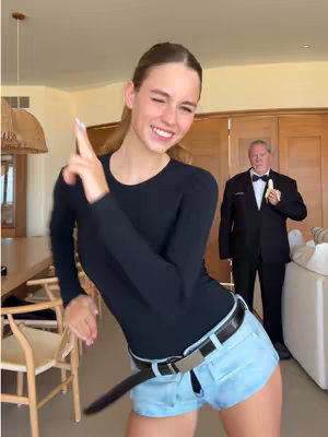 A young woman, smiling and pointing, strikes a pose in a living room setup, with a man holding a banana in the background.