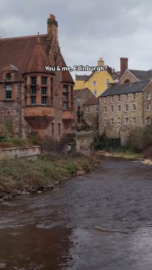 An eye-level shot captures a scenic view of Edinburgh, Scotland. A dark river flows toward the viewer, bordered by lush green vegetation. On the waterway's edge, a combination of tan and brown stone buildings line the banks. The buildings feature a mix of architectural styles, creating a picturesque cityscape under an overcast sky.