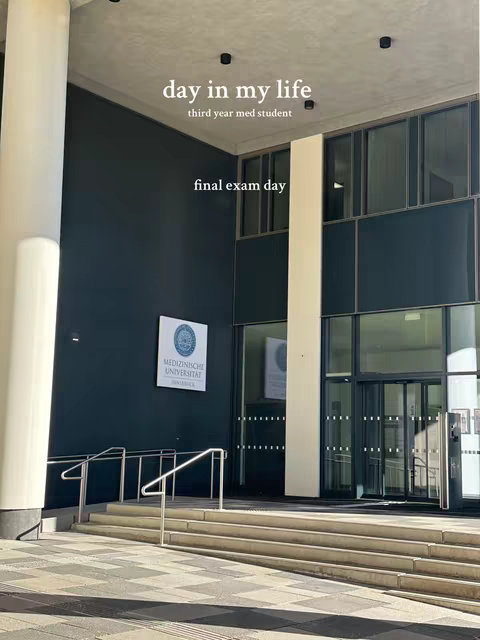 An exterior shot of a modern building featuring a grey and dark blue facade with large glass windows. Steps lead up to the entrance, and the sky is clear and bright. Text overlays indicate it is a 'Day in my life' video about a third-year med student on their final exam day.