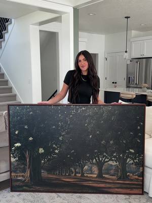 A woman with long dark hair stands in a well-lit living room. She is holding a large, framed painting that depicts a tree-lined road scene. She is smiling and looking at the camera. The room has white walls, a light-colored rug, and a staircase in the background.