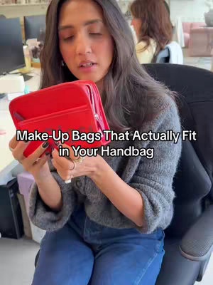 A woman with long dark hair holds a red makeup bag open, showcasing its contents and features. She is wearing a gray sweater and blue jeans and is seated. The background suggests an office setting, with a computer screen and another person partially visible.