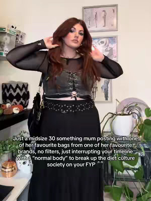 A woman with reddish-brown hair poses in a room with various decorations. She is wearing a black mesh long-sleeve top over a black bodice with a lace-up detail, and a black skirt. She has a shoulder bag and is holding her hands near her head. The background includes framed artwork and plants.
