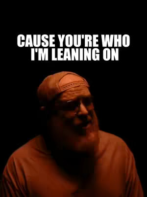 The image is a selfie-style shot featuring a man with a beard and a baseball cap against a black background. Text overlay displays the phrase "CAUSE YOU'RE WHO I'M LEANING ON" in large, white, bold font. The man is in the frame, partially lit with a reddish-orange hue.