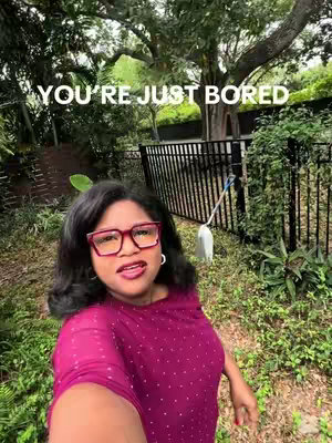 A selfie-style shot with a woman in the foreground, posing in a garden setting. She wears a dark pink top and large, red-framed glasses. The background includes greenery, a black fence, and a tall tree. The text overlay reads 'YOU'RE JUST BORED' in white block letters.