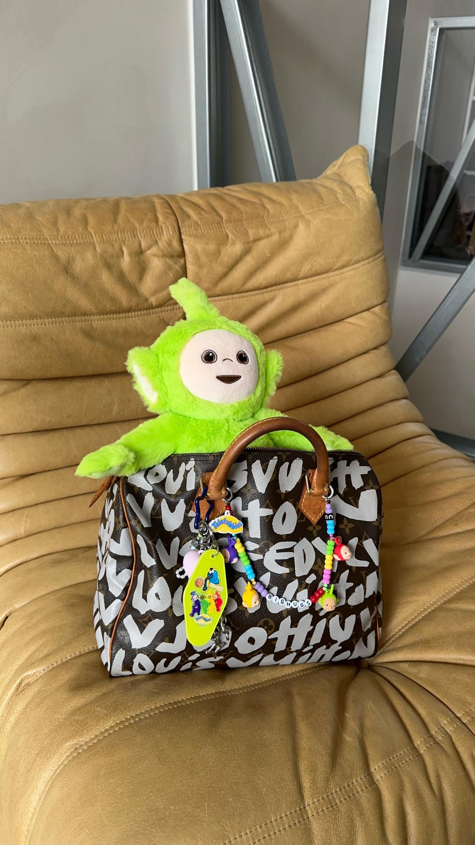 A green Teletubbies plush toy is placed on a Louis Vuitton handbag. The bag sits on a tan leather chair. There are charms hanging from the handle of the bag. The background includes metal support beams.