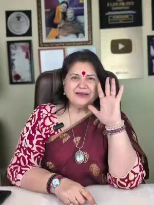 A middle-aged woman with black hair, wearing a maroon embroidered top and a necklace with a pendant. She's holding up her right hand with five fingers extended, as if counting or making a point. The background includes framed pictures, a YouTube logo, and a nondescript tan wall with a large picture frame.