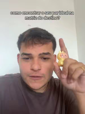 A man with short brown hair looking at the camera, holding a yellow cartoon character figurine in his right hand. A text overlay at the top says, "como encontrar o seu par ideal na matriz do destino?" The background is a plain white wall.
