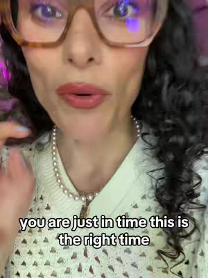 A close-up shot focuses on a woman's face. She wears large, square-framed glasses, and her lips are colored with red lipstick. A pearl necklace adorns her neck, topping a detailed white sweater. The background is blurred, suggesting an indoor setting possibly illuminated by pink and purple lighting.