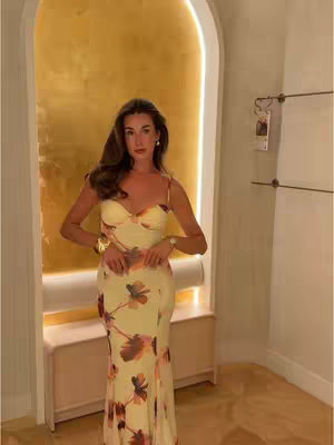 A woman in an elegant, floral-print dress stands indoors, posing in front of a gold accent wall within what appears to be a dressing room or boutique. She's framed by an archway and the warm backdrop creates a luxurious feel. She's wearing a watch and gold accessories.