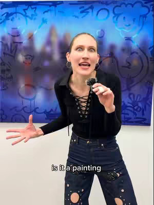 A woman with fair skin stands in front of a colorful abstract painting. She's wearing a black top, dark jeans with cutouts, and holding a microphone. The background is a blurred blue painting with abstract shapes.