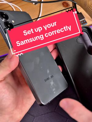The image shows a person holding a gray Samsung phone in both hands, with another phone and case visible in the background. Overlayed on the image is a red rectangle with the text "Set up your Samsung correctly" in white. The setting appears to be a desk or table with a wooden surface and a USB cable.