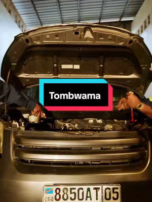 The cover image shows the front of a car with the hood open, revealing the engine. Two people are working on the engine. A rectangular text overlay with the word 'Tombwama' is in the center. The overall composition is a medium shot with a focus on the car's engine.