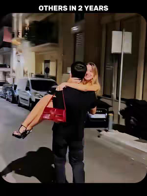 A man carries a smiling woman in his arms down a street at night. The background shows buildings and parked cars. The woman is wearing a black dress and red purse.