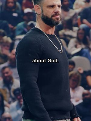 A man with a beard, wearing a black sweater and a silver necklace is the main focus. He stands against a blurred background of an auditorium. Text overlayed in the image says: 'about God.'