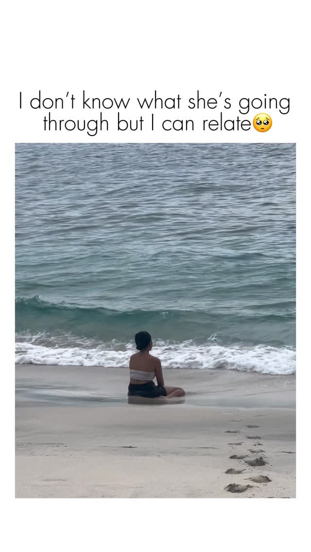 A person, seen from behind, is sitting cross-legged on a sandy beach near the water's edge. The ocean waves gently lap the shore. Text is overlaid at the top of the image.