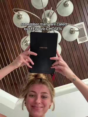 A woman, centered in the frame, smiling into the camera and holding a closed black book above her head. The book appears to be a Bible. The background shows a decorative light fixture with multiple bulbs and a wooden ceiling.