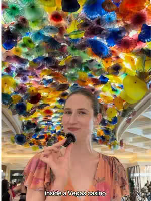 A vertical image features a woman standing in front of a vibrant, colorful glass sculpture that covers the ceiling. The sculpture is composed of various shapes and hues, creating a dynamic and artistic backdrop. The woman is the primary focus, dressed in a light-colored dress with floral patterns. She holds a dark-colored object towards the camera. The text overlay at the bottom of the image reads 'inside a Vegas casino'. The overall composition is well-lit, with a slightly blurred background hinting at the casino environment.