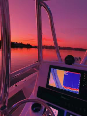 The image is taken from inside a boat, looking out at a sunset. The sky is a gradient of pink and orange, reflecting on the water. The boat's console and steering wheel are visible in the foreground, creating a contrast between the interior and the natural landscape.