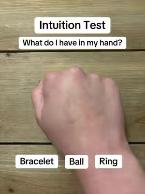 A close-up shot of a hand making a fist, likely belonging to a woman, with the back of the hand facing the camera. Above the hand, text boxes read 'Intuition Test' and 'What do I have in my hand?'. Below the hand are three text boxes with the words 'Bracelet,' 'Ball,' and 'Ring.' The background is a wooden surface.