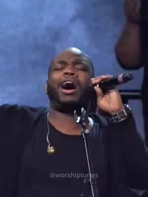 Image shows a close-up of a Black man singing into a microphone. He is wearing a black top and a watch. The background is a blurred blue, presumably a stage. The man's eyes are closed, and his mouth is open as if singing with great passion.