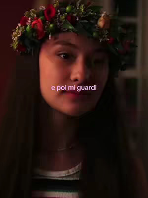 A medium shot showing a young woman wearing a floral crown, looking towards the camera. She appears to be inside, with warm lighting, and the text "e poi mi guardi" is overlaid towards the center of the frame.