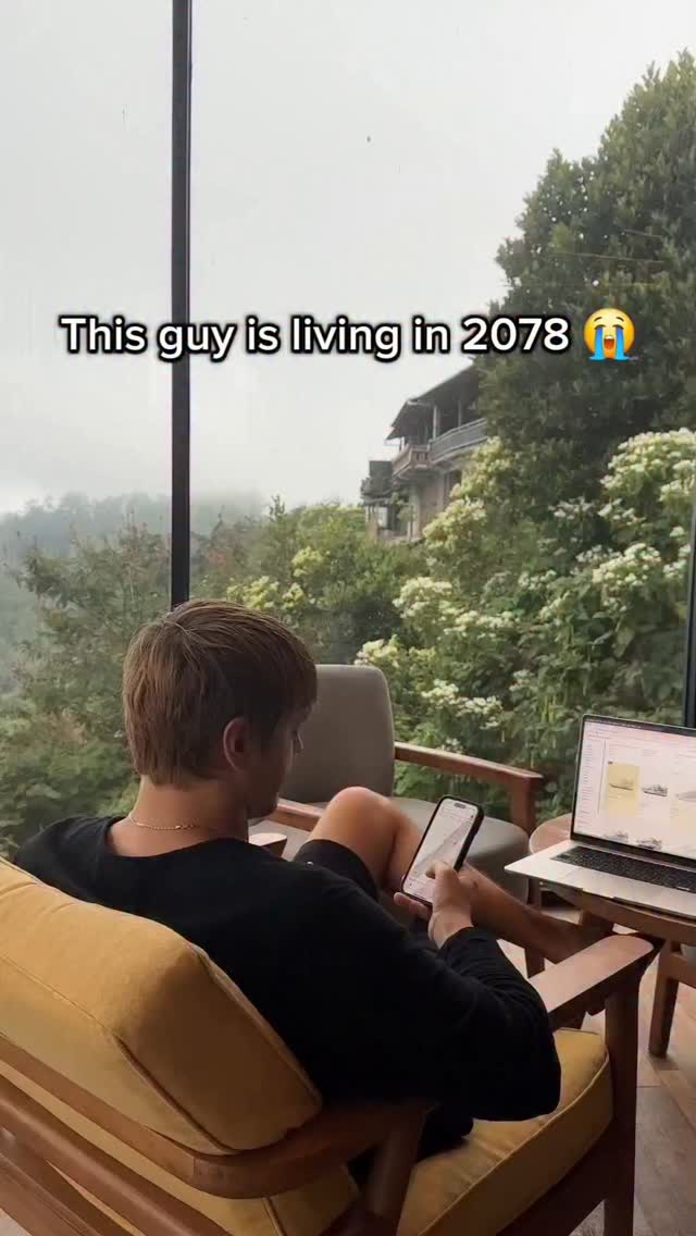A man is sitting in a chair with a laptop and a phone in a scenic outdoor setting. The background reveals a lush green landscape, with a mountainous or hilly terrain. The overall impression is modern and relaxed. Overlay text says, 'This guy is living in 2078' with a crying emoji.