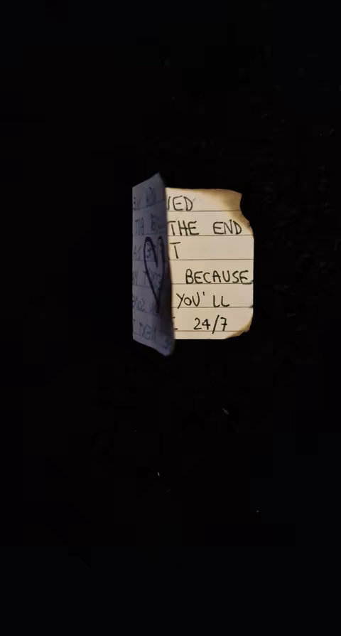 A close-up of a piece of paper with handwritten text, set against a stark black background. The paper appears to be torn and slightly burned at one edge giving an aged look and adding to the distressed theme. A simple heart drawing is present, symbolizing love or affection as well as a message that seems to suggest a relationship.