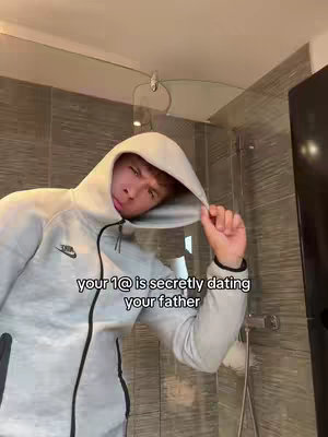A young man is in a tiled bathroom, wearing a gray hoodie. He's pulling the hood over his head with one hand. Text overlayed on the image reads, 'your 1@ is secretly dating your father.'