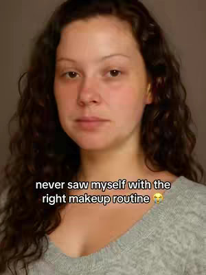 A woman with wavy, dark hair is centered in the frame, looking directly at the camera with a neutral expression. She appears to be makeup-free or with minimal makeup, as her complexion is natural. Her hair falls around her shoulders, and she wears a plain gray sweater against a plain background. Text overlays are present at the bottom, with the central focus on her face.