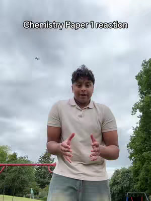 A young man stands outdoors facing the camera, hands gesturing outwards. The background features trees, a cloudy sky, and playground equipment. Text overlay at the top reads: Chemistry Paper 1 reaction.