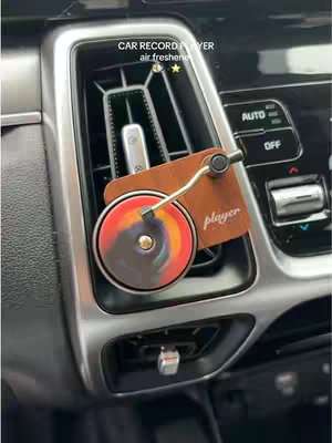 An air vent in a car with a custom air freshener shaped like a retro record player installed. The player has a brown wooden face, a silver arm, and a colorful vinyl record in the center. The car’s vent system is visible in the background with the controls and surrounding trim.