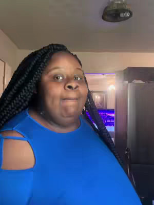 A medium shot shows a Black woman with long, braided hair, looking directly at the camera with a slightly surprised expression. She wears a blue top with cut-out shoulder details. The image is brightly lit, and the background includes glimpses of a room with a light fixture on the ceiling and a dark-colored object on the right.
