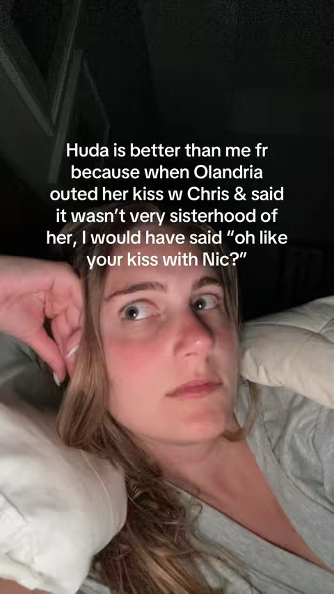A selfie-style shot featuring a young woman in a grey top, lying in bed with white bedding visible. She has light-colored hair, and a neutral expression, partially obscured by the frame's edge. Text is overlaid displaying a caption that appears to be a reaction to something.