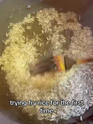 A top-down shot of rice being fried in a pan. The rice is glistening in oil, and a spatula is visible, stirring the rice. The text overlay says, "trying fry rice for the first time."