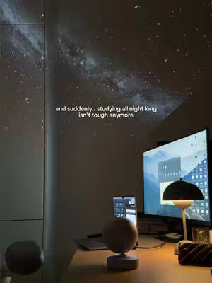 A dimly lit room with a desk and a galaxy projector. The image captures a desk setup in a dark room. The wall is illuminated by a projection of a starry night, suggesting use of a galaxy projector. The desk is cluttered with a laptop, a desk lamp, a monitor, and other stationery items. The overall impression is of a cozy, dimly lit study space.