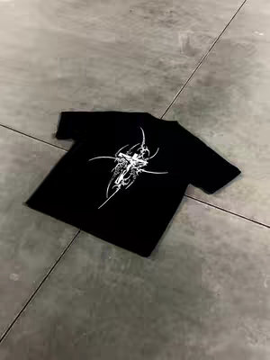 A black t-shirt lies diagonally across a gray concrete surface. The t-shirt has a white, abstract cross design on the front. The angle of the shot provides a top-down view.