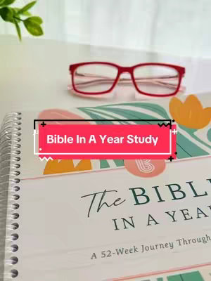 The image shows a hardcover book titled "The Bible in a Year" with a pink and floral design. Red-framed glasses are perched on top. The book is on a white surface, and a few leaves are visible in the upper left corner. A rectangular text overlay with the words "Bible In A Year Study" is on the book.