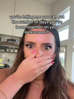 A woman with long brown hair is shown in a close-up shot, covering her mouth with her right hand. She has noticeable eye makeup and pink nail polish. The background suggests a room setting with some shelves and a window visible.